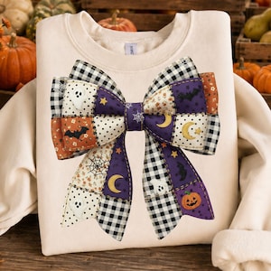 May include: Cream-colored sweatshirt featuring a large, patchwork bow design. The bow incorporates Halloween-themed patterns, including ghosts, bats, pumpkins, moons, stars, and spiderwebs. The bow's colors include black, white, orange, and purple.