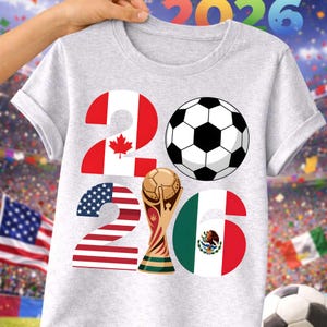 World Soccer Cup 2026 soccer PNG displayed on a light gray t-shirt mockup with a stadium background, featuring bold 2026 typography filled with USA, Canada, and Mexico flag designs, a soccer ball icon, and a gold trophy illustration. This high-quality transparent PNG is ideal for print on demand t-shirts, kids soccer shirts, fan apparel, sublimation projects, and football merchandise celebrating the 2026 world soccer tournament.