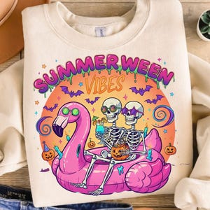 May include: Cream-coloured sweatshirt with a colourful graphic. The graphic features two skeletons wearing sunglasses, lounging on a pink flamingo float, and holding cocktails. The text "SUMMERWEEN VIBES" is above the skeletons, with bats and pumpkins.