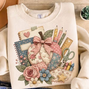 May include: A cream-colored crewneck sweatshirt featuring a graphic print of a quilted pumpkin with a pink bow, surrounded by school supplies including notebooks, pencils, a ruler, scissors, crayons, and floral accents in shades of pink and blue.