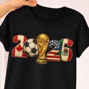 Puede incluir: Camiseta negra con el año "2026", un balón de fútbol y un trofeo dorado. Los números están diseñados con las banderas de Canadá, Estados Unidos y México.