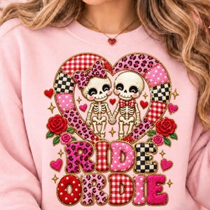May include: A light pink sweatshirt featuring a graphic of two skeletons holding hands inside a heart-shaped frame. The design includes red roses, small hearts, and the text RIDE OR DIE in a mix of leopard, plaid, and polka dot patterns.