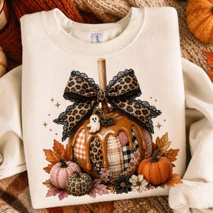 May include: A cream-colored crewneck sweatshirt featuring a graphic of a patchwork pumpkin with caramel-colored glaze. The pumpkin is topped with a large leopard print bow, a small ghost charm, and a bat charm, surrounded by autumn leaves and flowers.