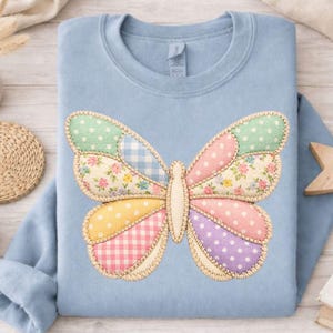 Może przedstawiać: Jasnoniebieska bluza z dużym motylem. Motyl składa się z patchworkowych sekcji z wzorami takimi jak kropki, kratka vichy i nadruki kwiatowe w pastelowych odcieniach. Krawędzie motyla są obszyte beżową nicią.