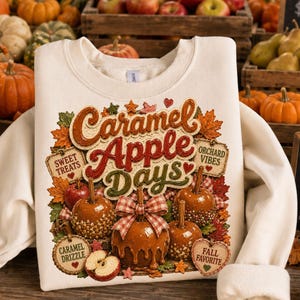 May include: Cream-colored sweatshirt featuring a graphic design with the text "Caramel Apple Days." The design includes caramel apples, fall leaves, and the phrases "Sweet Treats," "Orchard Vibes," "Caramel Drizzle," and "Fall Favorite."