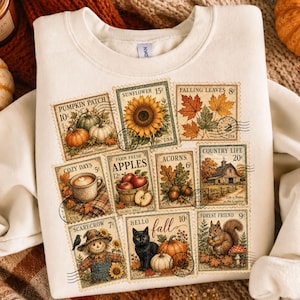 May include: A cream-coloured crewneck sweatshirt featuring a graphic print of nine vintage-style postage stamps. The stamps depict autumn themes including pumpkins, a sunflower, falling leaves, a coffee mug, apples, acorns, a barn, a scarecrow, a black cat, and a squirrel.