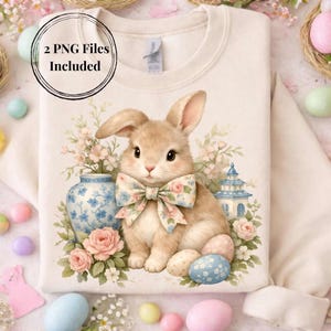 Puede incluir: Sudadera color crema con una ilustración de acuarela de un conejito con una pajarita floral, rodeado de flores, huevos y un jarrón azul y blanco. El texto "2 PNG Files Included" está en un círculo en la parte superior.