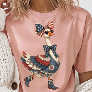 Puede incluir: Camiseta rosa con una ilustración de un ganso patriótico. El ganso lleva un lazo, gafas de sol y un peto vaquero con una combinación de colores rojo, blanco y azul. El ganso también lleva botas con estrellas.