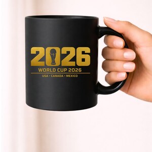 Black coffee mug mockup showing a gold soccer design featuring the year 2026 with a stylized trophy icon forming the zero and text reading World Cup 2026 USA Canada Mexico. This 2026 sports design is ideal for sublimation printing on mugs, tumblers and drinkware as well as apparel designs for soccer fans. Perfect digital graphic for print on demand sellers and Cricut creators producing sports themed merchandise.