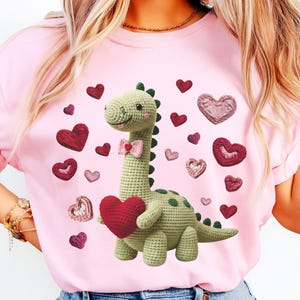 May include: Pink t-shirt featuring a crocheted dinosaur holding a red heart, surrounded by various heart shapes. The dinosaur is light green with a pink bow tie and green spots. The hearts are in shades of red and pink.