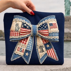 May include: A navy blue decorative pillow featuring a large bow design. The bow is made of fabric patches with patriotic themes, including the American flag, Statue of Liberty, and the text "USA". The bow is trimmed with lace.