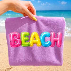 May include: A lavender beach towel with the word "BEACH" in colorful, bubble-style letters. The letters are pink, green, yellow, blue, and pink. The towel is held up against a background of a sandy beach and ocean.
