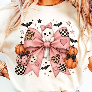 May include: A cream-colored t-shirt featuring a graphic of a white ghost with a pink bow, surrounded by orange and pink pumpkins, black bats, and stars. The design includes leopard print and checkered patterns on the bow and pumpkin accents.