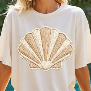 Może przedstawiać: Kremowy t-shirt z haftowanym wzorem muszli. Muszla jest w odcieniach beżu i kremu, o teksturowanym wyglądzie. Koszulka ma krótkie rękawy i okrągły dekolt.