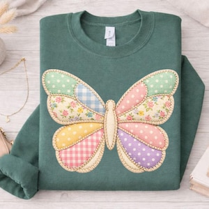 May include: A dark teal sweatshirt featuring a large, appliqued butterfly. The butterfly's wings are composed of various patterned fabrics, including polka dots, floral prints, and gingham in pastel colors. The edges of the butterfly are stitched with a decorative thread.