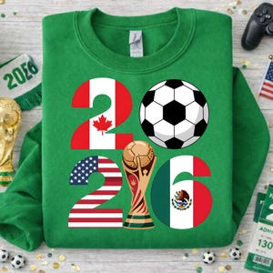 World Soccer Cup 2026 inspired PNG design displayed on a green sweatshirt mockup, showing the year 2026 with United States, Canada, and Mexico flag details, a black and white soccer ball, and a golden trophy graphic. This transparent PNG file is perfect for print on demand apparel, kids soccer sweatshirts, fan merchandise, sublimation printing, and football-themed clothing designs for the 2026 international soccer event.
