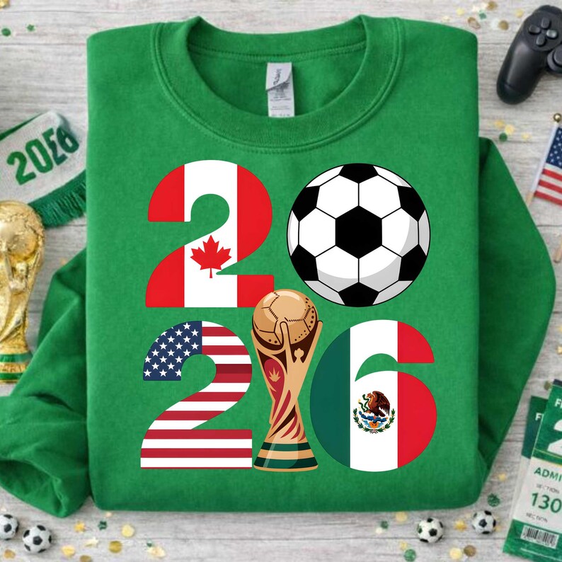 World Soccer Cup 2026 inspired PNG design displayed on a green sweatshirt mockup, showing the year 2026 with United States, Canada, and Mexico flag details, a black and white soccer ball, and a golden trophy graphic. This transparent PNG file is perfect for print on demand apparel, kids soccer sweatshirts, fan merchandise, sublimation printing, and football-themed clothing designs for the 2026 international soccer event.