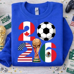 World Soccer Cup 2026 soccer PNG shown on a royal blue sweatshirt mockup, featuring the year 2026 designed with USA, Canada, and Mexico flag elements, a classic soccer ball, and a gold trophy illustration. This high-resolution transparent PNG is ideal for print on demand sweatshirts, youth soccer apparel, fan gear, sublimation projects, and custom football shirt designs celebrating the 2026 world soccer tournament hosted in North America.