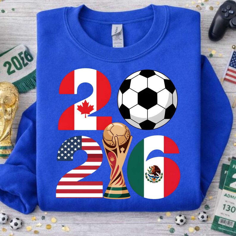 World Soccer Cup 2026 soccer PNG shown on a royal blue sweatshirt mockup, featuring the year 2026 designed with USA, Canada, and Mexico flag elements, a classic soccer ball, and a gold trophy illustration. This high-resolution transparent PNG is ideal for print on demand sweatshirts, youth soccer apparel, fan gear, sublimation projects, and custom football shirt designs celebrating the 2026 world soccer tournament hosted in North America.