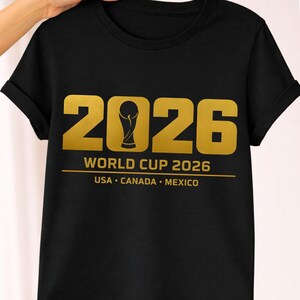 Black shirt mockup presenting a bold gold 2026 soccer PNG design created for soccer fan shirts and sports merchandise. The graphic displays large varsity style numbers with a stylized trophy icon forming the zero and text reading Soccer Cup 2026 USA Canada Mexico. This sports design is perfect for sublimation printing, Cricut and Silhouette machines, print on demand apparel and custom soccer merchandise celebrating the upcoming 2026 global tournament.