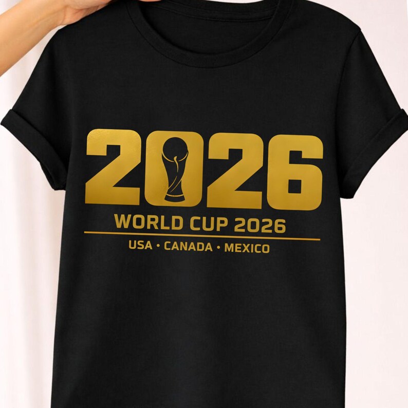 Black shirt mockup presenting a bold gold 2026 soccer PNG design created for soccer fan shirts and sports merchandise. The graphic displays large varsity style numbers with a stylized trophy icon forming the zero and text reading Soccer Cup 2026 USA Canada Mexico. This sports design is perfect for sublimation printing, Cricut and Silhouette machines, print on demand apparel and custom soccer merchandise celebrating the upcoming 2026 global tournament.