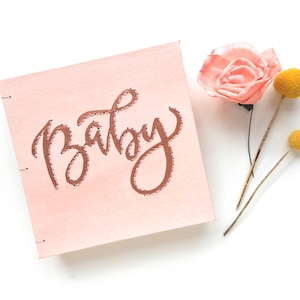 Puede incluir: Un libro rosa con la palabra "Baby" bordada en hilo de cobre. El libro está rodeado de una rosa rosa y dos pompones amarillos.