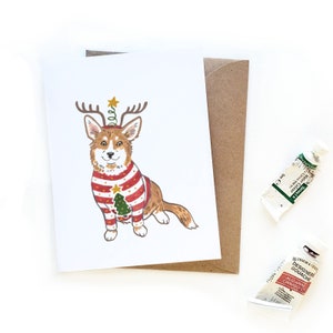 Może przedstawiać: Kartka świąteczna z kreskówkową ilustracją corgi w czerwono-białej, pasiastych swetrze z małym drzewem bożonarodzeniowym. Corgi ma poroże i gwiazdę na głowie.