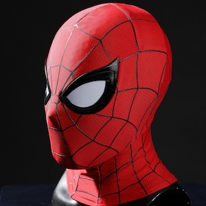 May include: A red fabric mask featuring a black web pattern across the surface. The mask includes large, angular white eye lenses framed by thick black borders. The design covers the entire head and neck area, displaying a textured material finish.