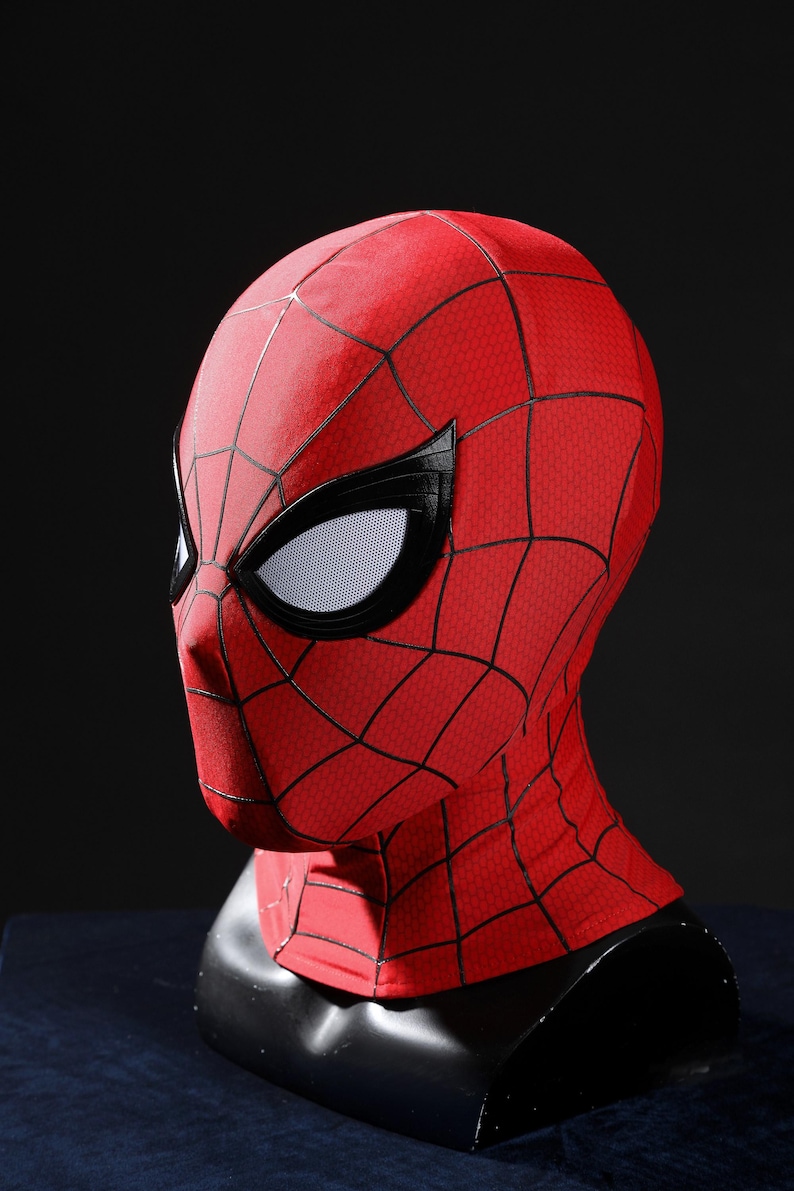 May include: A red fabric mask featuring a black web pattern across the surface. The mask includes large, angular white eye lenses framed by thick black borders. The design covers the entire head and neck area, displaying a textured material finish.