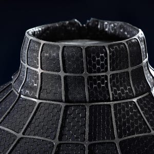 K&ouml;nnte beinhalten: Ein schwarzes Kleidungsst&uuml;ck mit hohem Kragen und einem erhabenen Gittermuster auf der Oberfl&auml;che. Der Stoff hat eine texturierte, wabenartige Oberfl&auml;che in jedem Gitterquadrat, und der Kragen ist mit einem markanten, dicken, dunkelgrauen Netzdesign strukturiert.