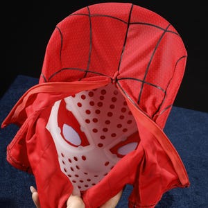 May include: A red fabric mask featuring a black web pattern and a zipper closure. The mask includes a white plastic inner shell with circular ventilation holes and white mesh eye coverings, designed to maintain the shape of the head covering.