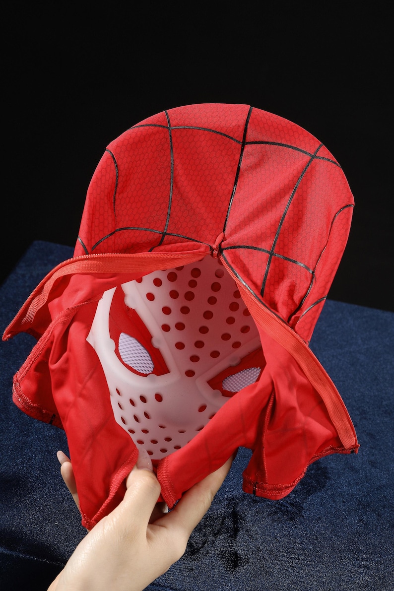 May include: A red fabric mask featuring a black web pattern and a zipper closure. The mask includes a white plastic inner shell with circular ventilation holes and white mesh eye coverings, designed to maintain the shape of the head covering.