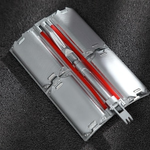 May include: A rectangular silver-colored metal mold featuring four distinct recessed sections and a central channel. A bright red rod is positioned within the center of the mold, which includes a small, pronged metal attachment at the bottom end.