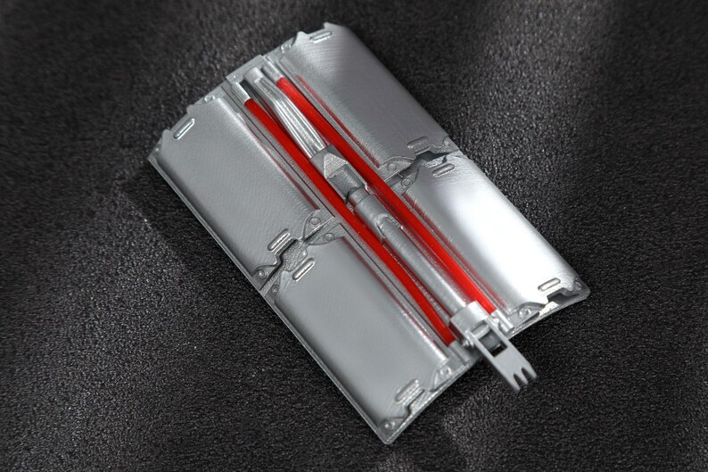 May include: A rectangular silver-colored metal mold featuring four distinct recessed sections and a central channel. A bright red rod is positioned within the center of the mold, which includes a small, pronged metal attachment at the bottom end.