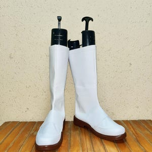 以下が含まれることがあります： 茶色のソールが付いた白いニーハイブーツのペア。ブーツは、丸いつま先と足の上部にストラップが付いたシンプルなデザインです。黒いブーツシェイパーがブーツの内側に挿入されています。