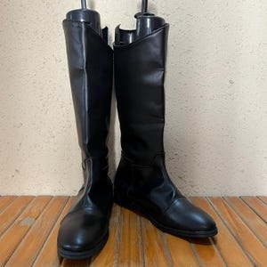 Może przedstawiać: Para czarnych butów do kolan. Buty wykonane są z gładkiego, błyszczącego materiału i mają prosty, klasyczny design. Są prezentowane na drewnianej powierzchni.