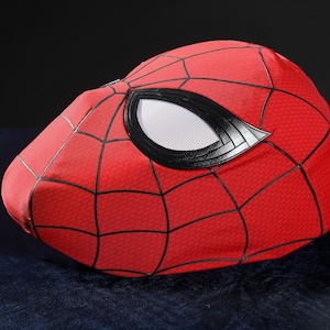 May include: A red fabric mask featuring a black web pattern across the surface. The mask includes large, almond-shaped eye openings framed by thick black borders, with white mesh lenses covering the eye areas to complete the design.