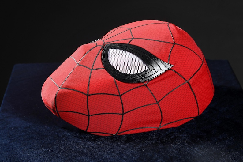 May include: A red fabric mask featuring a black web pattern across the surface. The mask includes large, almond-shaped eye openings framed by thick black borders, with white mesh lenses covering the eye areas to complete the design.