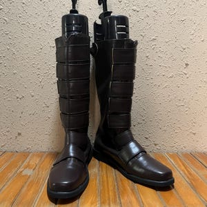 Puede incluir: Un par de botas marrones hasta la rodilla con múltiples correas y hebillas. Las botas tienen punta cuadrada y se exhiben sobre una superficie de madera. Diseño elegante y moderno.