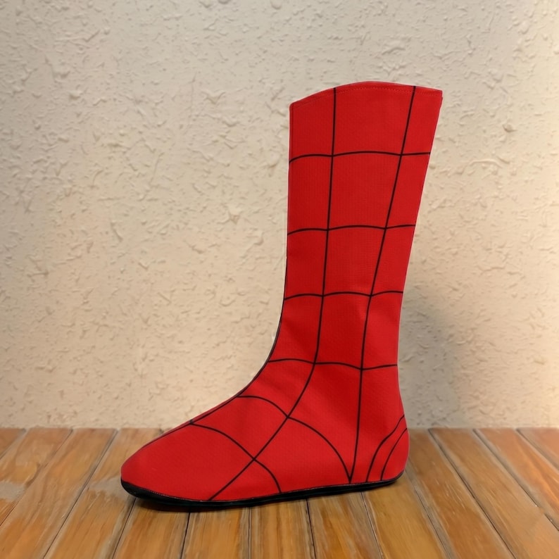Puede incluir: Una funda para bota de color rojo con un patr&oacute;n de cuadr&iacute;cula negra que se asemeja a una telara&ntilde;a. La tela es lisa y cubre toda el &aacute;rea del pie y el tobillo, extendi&eacute;ndose hacia arriba en una silueta alta y recta con punta redondeada y suela plana.