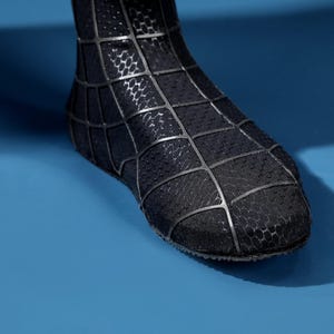 K&ouml;nnte beinhalten: Ein schwarzer Stiefel mit einer strukturierten, wabenartigen Oberfl&auml;che, die mit einem erhabenen, gitterartigen Netzmuster in mattem Schwarz &uuml;berzogen ist. Das Schuhwerk bedeckt den Fu&szlig;- und Kn&ouml;chelbereich und zeigt eine strukturierte Form.