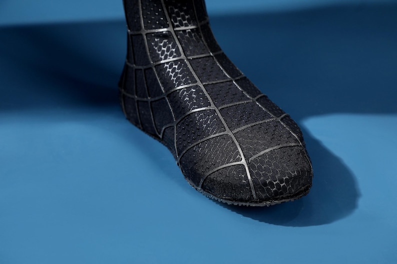 K&ouml;nnte beinhalten: Ein schwarzer Stiefel mit einer strukturierten, wabenartigen Oberfl&auml;che, die mit einem erhabenen, gitterartigen Netzmuster in mattem Schwarz &uuml;berzogen ist. Das Schuhwerk bedeckt den Fu&szlig;- und Kn&ouml;chelbereich und zeigt eine strukturierte Form.