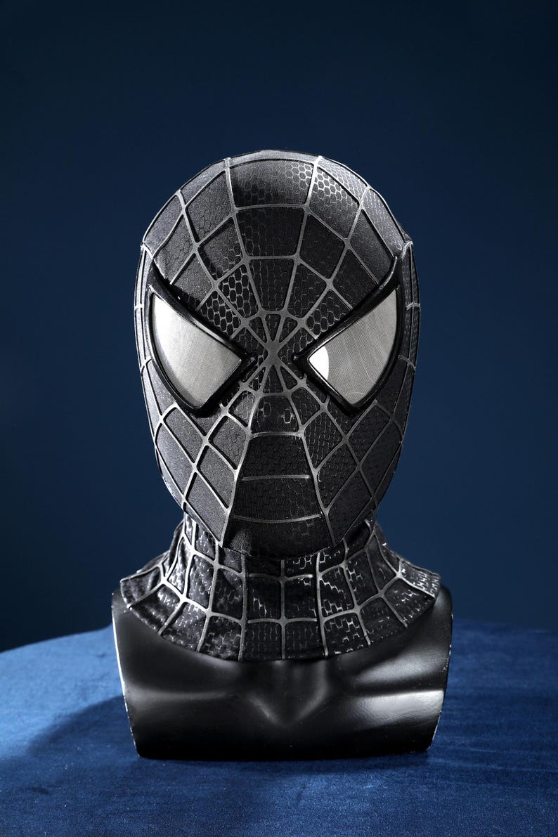 K&ouml;nnte beinhalten: Eine schwarze Vollkopfmaske mit texturiertem Wabenmuster und erhabenen silbernen Netzlinien. Die Maske verf&uuml;gt &uuml;ber zwei gro&szlig;e, kantige, silber umrandete Augenlinsen mit reflektierender Oberfl&auml;che, die den Kopf- und Halsbereich bedecken.