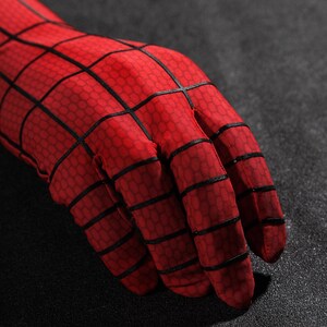 May include: A red fabric glove featuring a black grid pattern across the surface. The material has a subtle hexagonal texture throughout, and the black lines are raised, creating a distinct web-like design that covers the fingers and the wrist area.