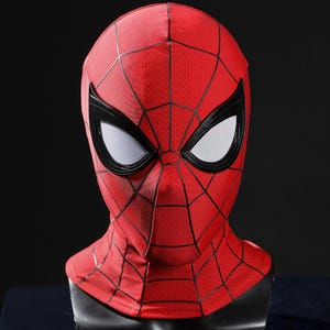 May include: A red fabric mask featuring a black web pattern across the surface. The mask includes large, almond-shaped eye openings framed in thick black borders with white mesh lenses, covering the entire head and neck area in a fitted design.