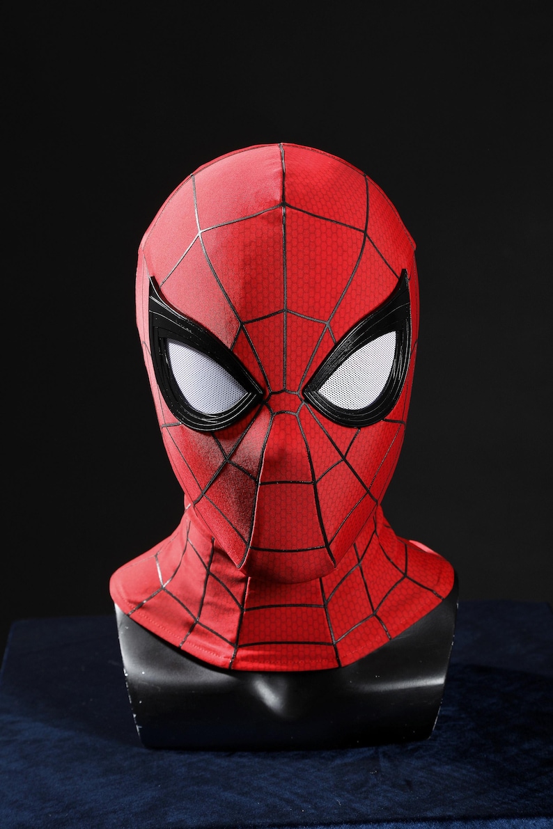 May include: A red fabric mask featuring a black web pattern across the surface. The mask includes large, almond-shaped eye openings framed in thick black borders with white mesh lenses, covering the entire head and neck area in a fitted design.