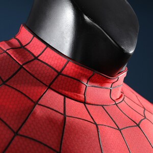 May include: A red fabric garment featuring a black web pattern with a high mock neck collar. The material displays a subtle hexagonal texture throughout the surface, worn on a black mannequin torso against a dark blue background.