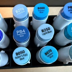 Könnte beinhalten: Eine Sammlung blauer Marker in einem schwarzen Organizer. Die Kappen sind in verschiedenen Blautönen gehalten und mit Namen wie "Indian Blue", "Vivid Blue" und "Crystal Blue" beschriftet. Die Marker sind zylindrisch mit weißen Körpern und blauen Kappen.