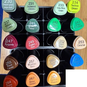 Op de afbeelding: Een verzameling kleurrijke stiften in een zwarte organizer. Elke stift heeft een kleurnaam en nummer, waaronder groentinten, rood, bruin en blauw. De stiften zijn in rijen gerangschikt en tonen een verscheidenheid aan tinten.