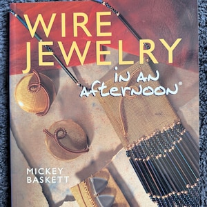 Könnte beinhalten: Ein Buch mit dem Titel "WIRE JEWELRY in AN Afternoon" von Mickey Baskett. Das Cover zeigt Beispiele für Drahtschmuck, darunter ein goldfarbenes gewebtes Stück mit schwarzen Perlen und Kupferdrahtdesigns. Der Hintergrund ist eine Mischung aus Rot und Beige.